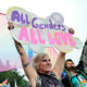 A person holds a sign that says "all genders, all love" with a pride flag flying in the background