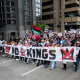 A large group of people walk through a street and hold a banner that says "No Kings"