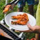 Outside by a lake, a man takes chicken drumsticks off of a grill onto a plate, kids surround him