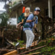 A person walks with a waking stick carrying a baby on their back outside through debris