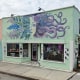 The exterior of Kilted Mermaid bar, a mint green painted building with a painting of a woman's face with plants and tentacles reaching out from either side