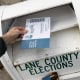 A hand hovers a ballot over a ballot drop box