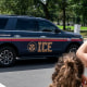 An Immigration and Customs Enforcement police SUV patrols on the road as people watch from the sidewalk