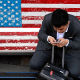 In this April 10, 2018, photo, a man on a bench with an American flag checks his smartphone in Beijing.