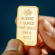 Two hands hold a small gold bar with imprinted words that read "Suisse 1 Ounce Fine Gold 999.9"