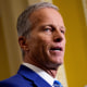 Senate Majority Leader John Thune raises his hand as he speaks to the press.