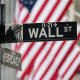 A street sign that shows the intersection of Wall Street and Broad Street, American flags are seen blurred in the background