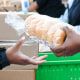 People in line at a food bank, a woman passes someone a bag of bagels from across a table lined with food