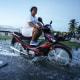 A woman rides her scooter through floodwaters next to a boy sitting near a plank on the flooded street