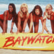 Actors pose with a large surfboard reading "Baywatch"