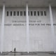A person walks a dog in front of the tall facade of a building with lettering reading THE DONALD J. TRUMP AND THE JOHN F. KENNEDY MEMORIAL CENTER FOR THE PERFORMING ARTS.