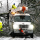 Two workers in blaze yellow suits work to clear fallen trees from a snow-covered roadway. 