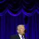 President Trump speaking into a microphone in front of a blue velvet stage backdrop.