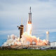 A rocket launches off a pad next to a building with large text on the front that reads "SpaceX"