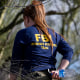 A woman with a long ponytail and a blue longsleeved shirt that says FBI EVIDENCE RESPONSE TEAM walks among trees.