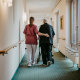 Rear view of female caregiver assisting senior woman walking with mobility walker in corridor
