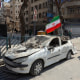 A man walks past an Iranian flag fluttering above the wreckage of a car.