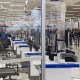 Empty TSA desks behind plastic barriers at an airport. A line of people is visible in the background.