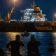 Two men are silhouetted against lights from large tankers floating across the water at night.