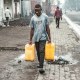 A man carries two yellow water jugs outside on the sidewalk