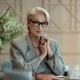 Meryl Streep clasps her hands while seated at a table wearing a suitcoat and black eyeglasses.