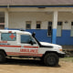 A white 4x4 with red lettering reading AMBULANCE parked on a dirt road outside a one-story concrete building.