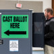 James Couch votes at a voting booth inside of a room, a large green sign that reads "cast ballot here" in black text is on the side of the voting booth
