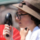 A woman wearing a hat holds an electric fan to cool her face while standing behind a metal barricade