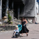 A mother pushes a stroller past a damaged building.