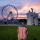 A man in a shirt with stars and stripes of the American flag stands in front of a Ferris Wheel at sunset.