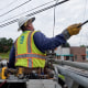 An AT&T technician works on a telephone pole, wearing a light green hazard vest with the AT&T logo and a white hard hat