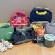Back-to-school gear for kids laid out on a wood floor: two backpacks (pink strawberry print and navy), a green lunchbox, white toddler sneakers, a folded jacket, Little Spoon snack pouches, a kids' smartwatch, an insulated food container, and a floral gingham lunch bag.