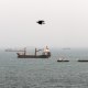 Numerous commercial vessels float under foggy skies as a black bird flys by in the foreground.
