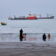 People stand by the beach where commercial vessels are seen in the horizon.