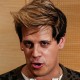 Milo Yiannopoulos speaks into microphones to reporters inside of a room, his shadow can be seen prominently on the wall behind him