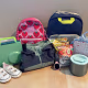 Back-to-school gear for kids laid out on a wood floor: two backpacks (pink strawberry print and navy), a green lunchbox, white toddler sneakers, a folded jacket, Little Spoon snack pouches, a kids' smartwatch, an insulated food container, and a floral gingham lunch bag.