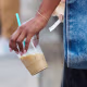 A guest wears a thin gold bracelet on the wrist, a large rectangular washed blue denim Telfar tote with dual top handles, a long shoulder strap, holding a clear plastic cup of iced coffee with a turquoise straw.