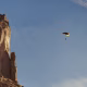 A person floating under a red, black and yellow parachute in front of a large stone cliff.