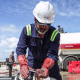 A worker in a hard hat twists the lever of a pipe outside near large trucks getting filled with oil