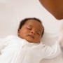 A Black baby dressed in a white onesie sleeps in a crib as a parent looks over.