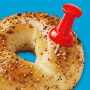 Photo illustration of an everything bagel with a red locator pin stuck in it.