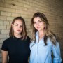 Lea Burbidge Izquierdo, left, and Francesca Albo stand next to each other in front of a beige brick wall