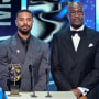 Michael B. Jordan and Delroy Lindo stand at a lectern on stage during an award ceremony. They look nonplussed.