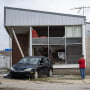 People stand outside of a damaged building with a damaged car parked out front