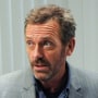 Hugh Laurie sits inside of a doctor's office next to medical supplies as he speaks to a patient sitting in front of him