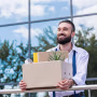 happy businessman with cardboard box with office supplies in hands standing outside office building, quitting job concept