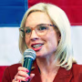Image: Ashley Hinson standing in front of a USA flag holding a microphone.